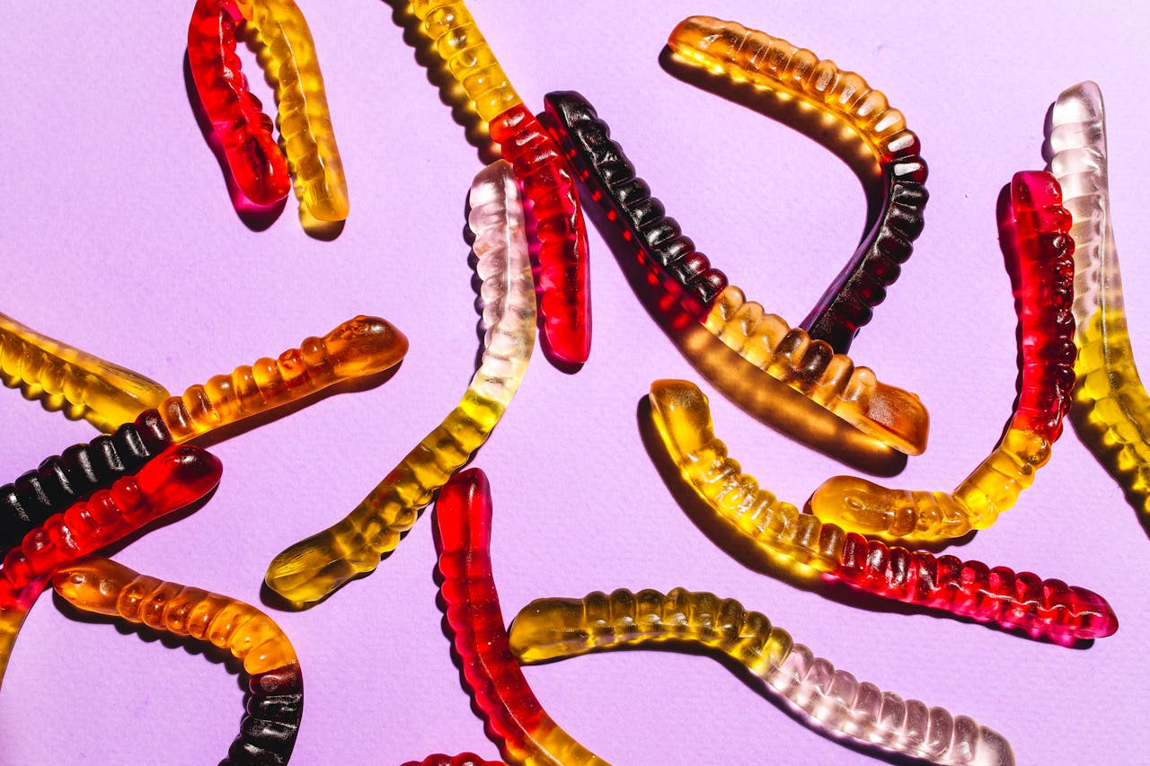 Colorful gummy worm candies, red, yellow, orange, black, arranged on a light purple background, translucent and chewy