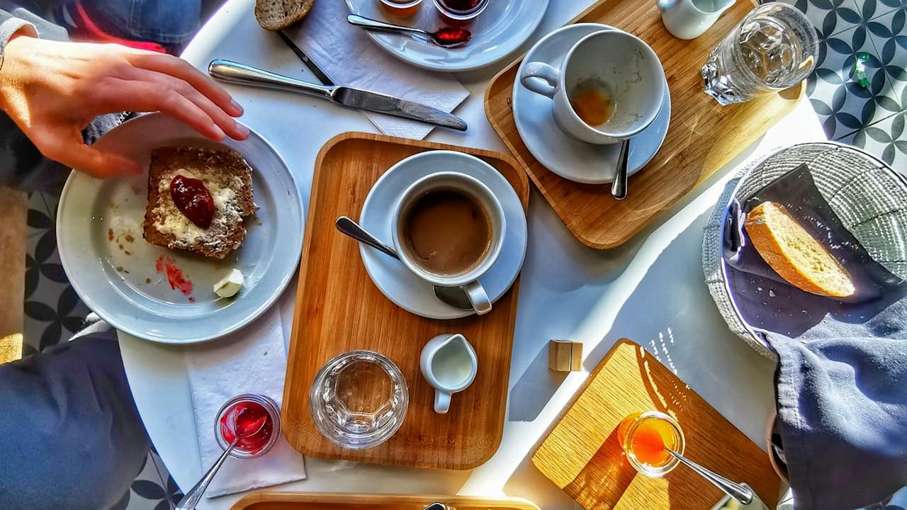 Breakfast table scene, cups of coffee partially drunk, small jars of jam, glasses of water, wooden serving trays, bread basket with remaining slice