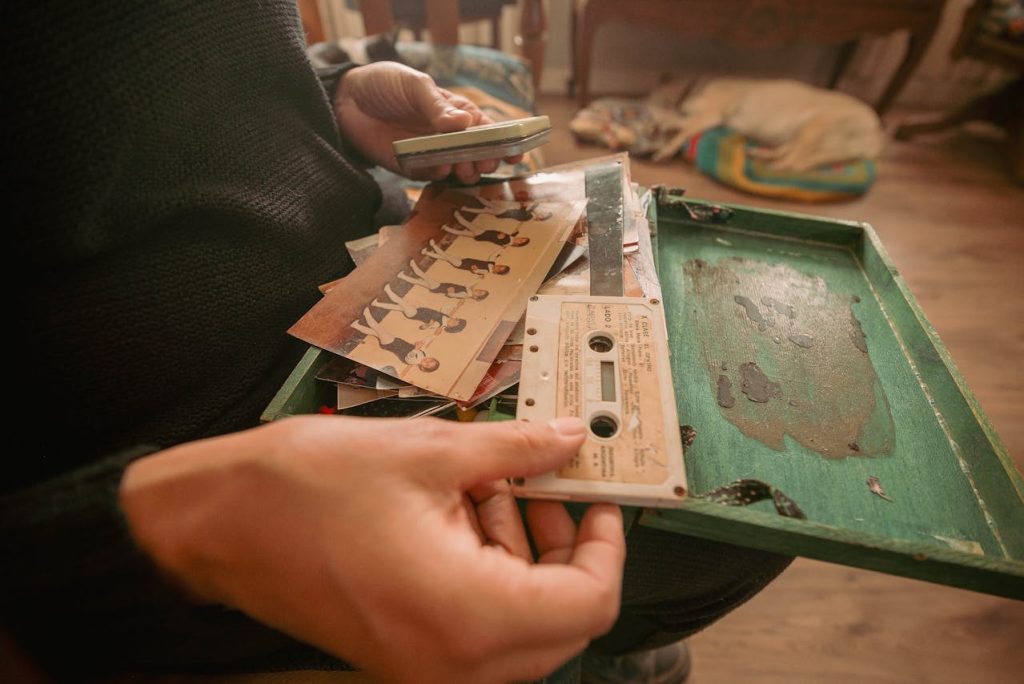 Person holding vintage cassette tapes, music tracklist cards, green box, cozy indoor setting, nostalgic atmosphere