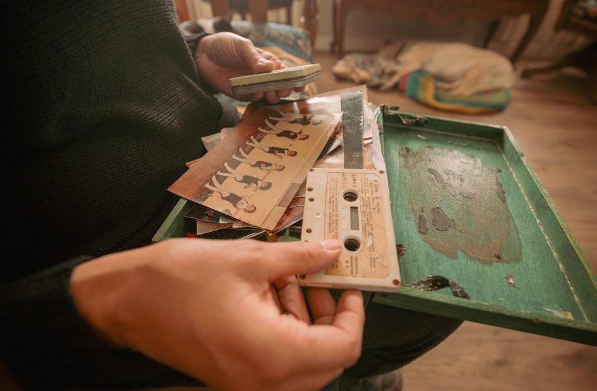 Person holding vintage cassette tapes, music tracklist cards, green box, cozy indoor setting, nostalgic atmosphere