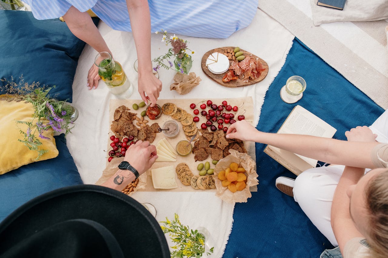 Indoor picnic setup with people reaching for snacks on a blanket surrounded by pillows, drinks, and flowers