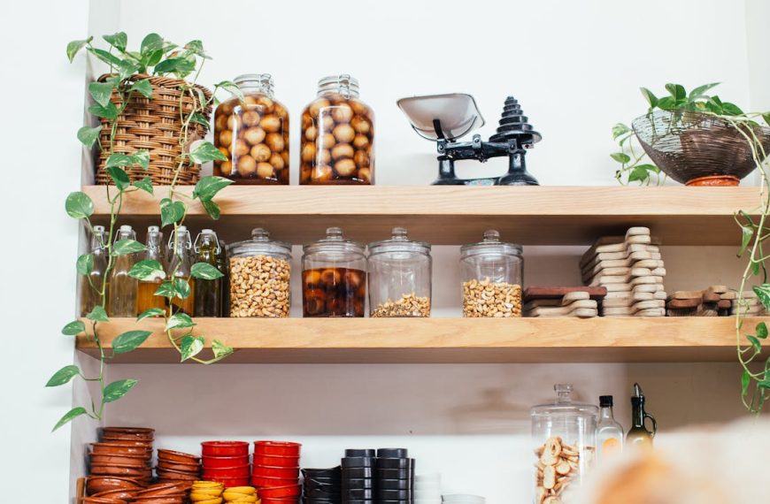 Kitchen pantry with wooden shelves, glass jars filled with nuts and preserved fruits, oil bottles, small bowls, cutting boards, green hanging plants