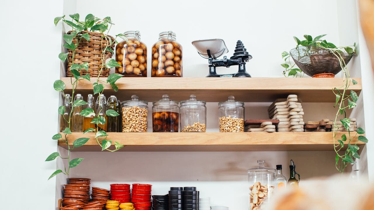Kitchen pantry with wooden shelves, glass jars filled with nuts and preserved fruits, oil bottles, small bowls, cutting boards, green hanging plants