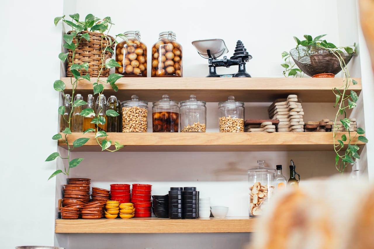 Kitchen pantry with wooden shelves, glass jars filled with nuts and preserved fruits, oil bottles, small bowls, cutting boards, green hanging plants