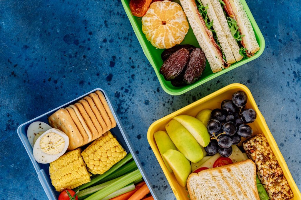 Three colorful lunch boxes on a blue surface, containing sandwiches, fruits, vegetables, boiled egg, crackers, granola bar