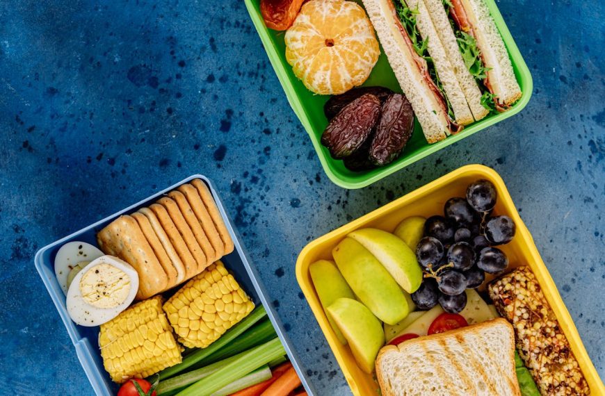 Three colorful lunch boxes on a blue surface, containing sandwiches, fruits, vegetables, boiled egg, crackers, granola bar