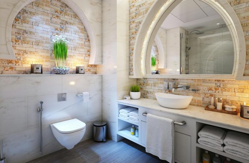 Modern bathroom with arched stone wall accents, wall-mounted toilet, vessel sink on white vanity, large mirror, decorative lighting, indoor plant on shelf