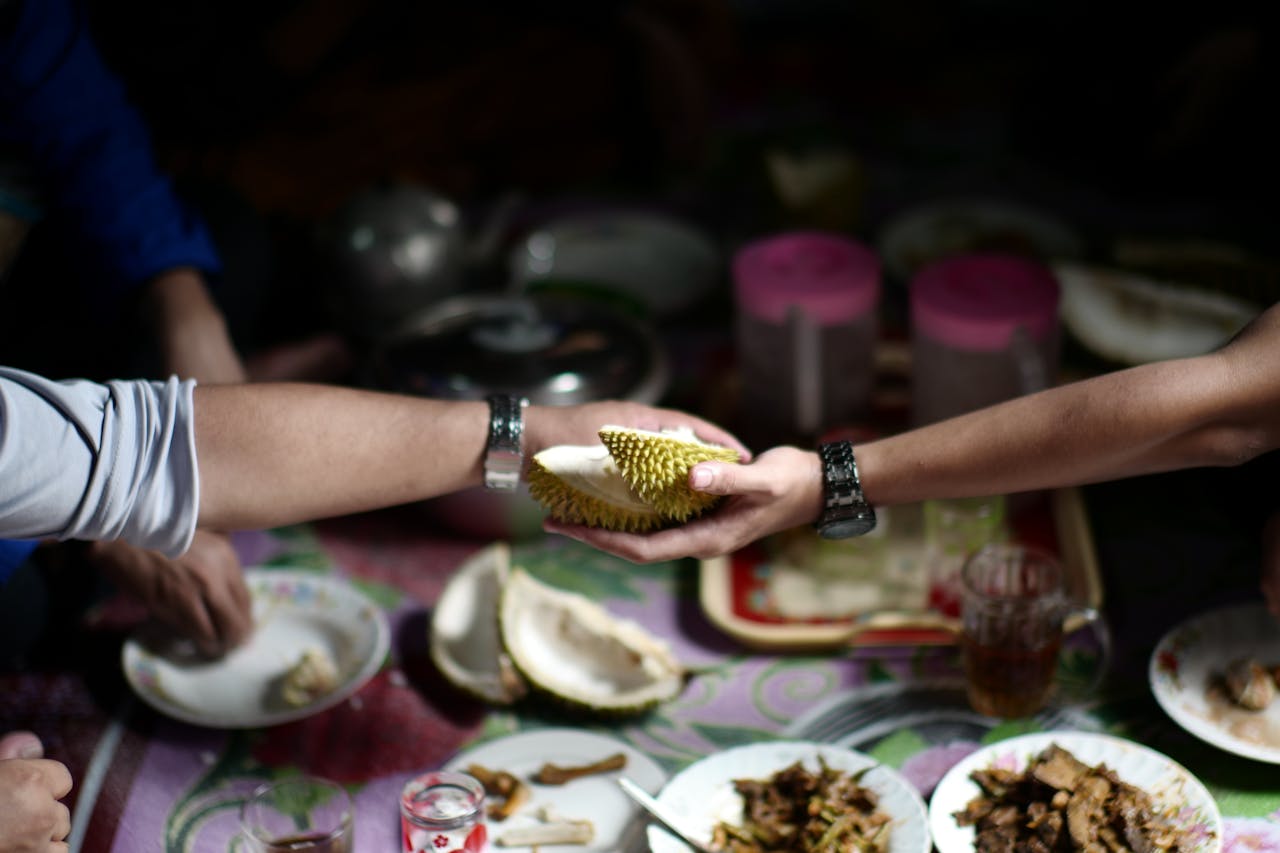 Two people exchanging a piece of durian, hands reaching across a table, plates of food and drinks in the background