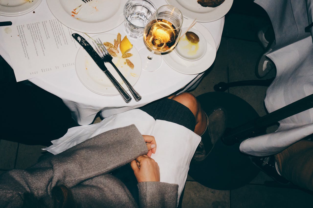 Overhead view of a dining table, white tablecloth, utensils placed on a plate, a menu on the table, person sitting with a white napkin on their lap