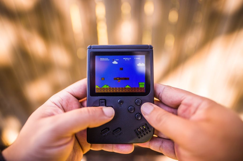 Close-up of hands holding black handheld retro gaming console, Super Mario level on screen, outdoor blurred background, directional pad and buttons visible