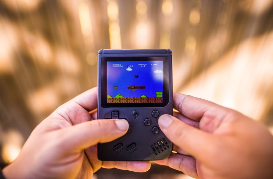 Close-up of hands holding black handheld retro gaming console, Super Mario level on screen, outdoor blurred background, directional pad and buttons visible