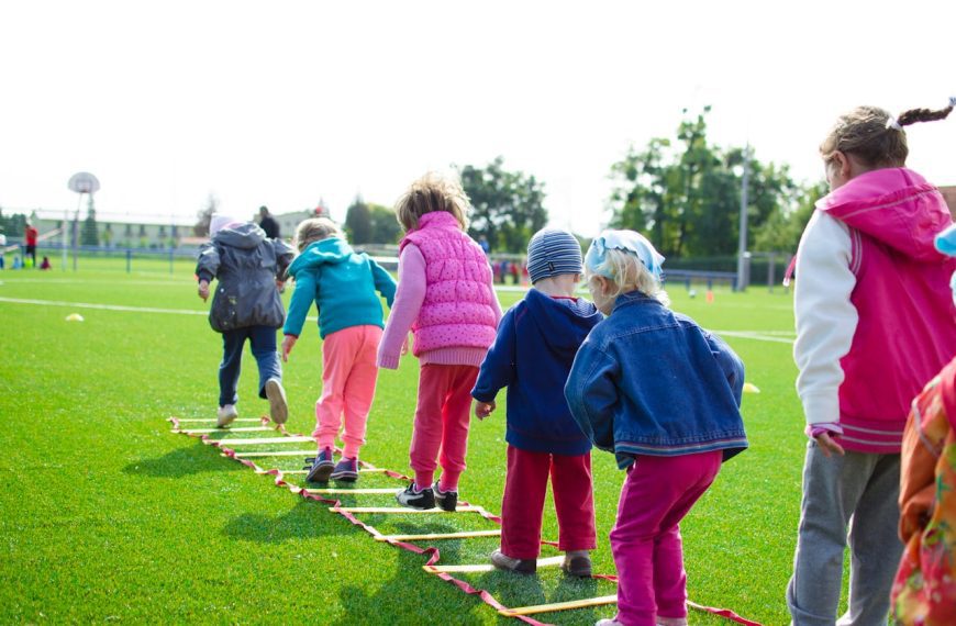Young children playing on a grassy field, walking in line on an agility ladder, dressed in colorful jackets and hats