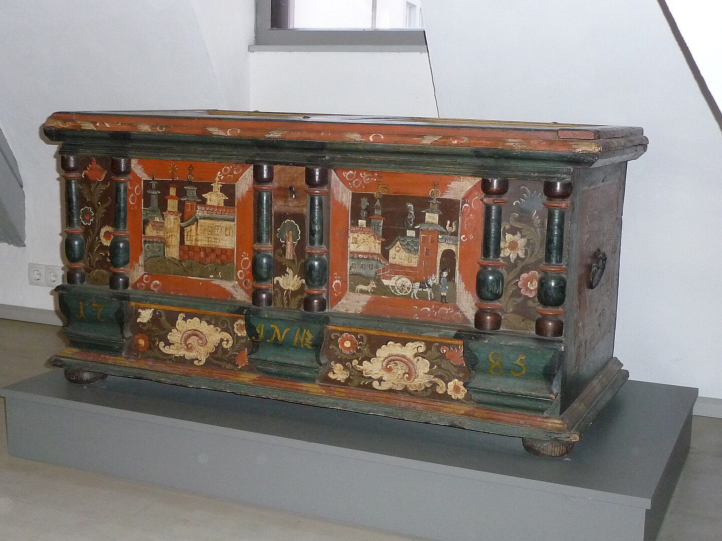 Antique hand-painted wooden chest or trunk with folk art decorations on terracotta-orange background. Features painted village scenes, floral motifs, turned columns, and dated "1785." Displayed on gray platform in museum setting with white walls