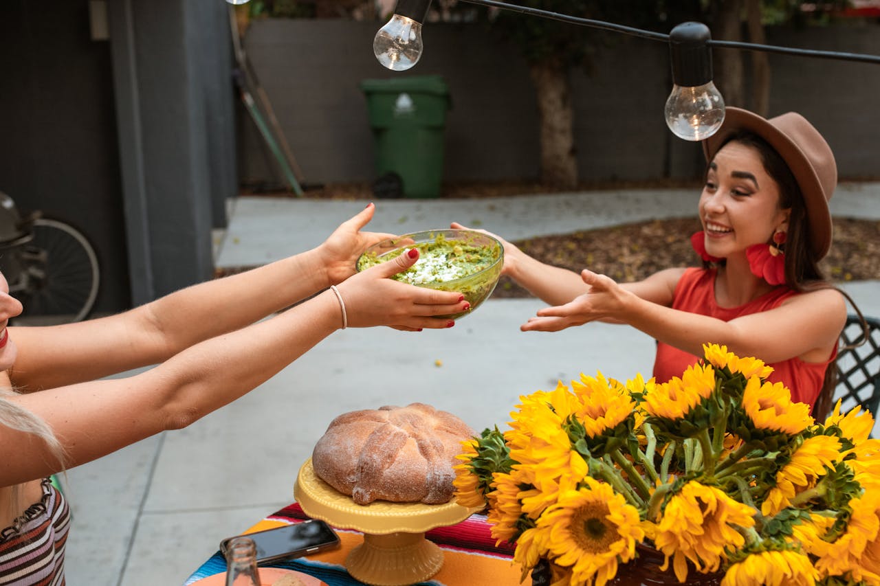 Two women outdoors passing a bowl of guacamole with cheese, a table decorated with sunflowers, a loaf of bread, string lights hanging above, casual and festive atmosphere