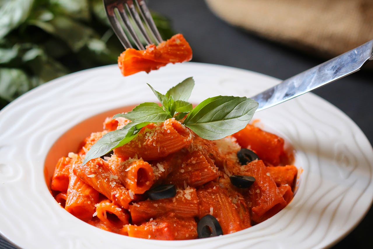 Bowl of rigatoni pasta with red sauce, garnished with basil leaves, black olives, and grated cheese, fork lifting a bite