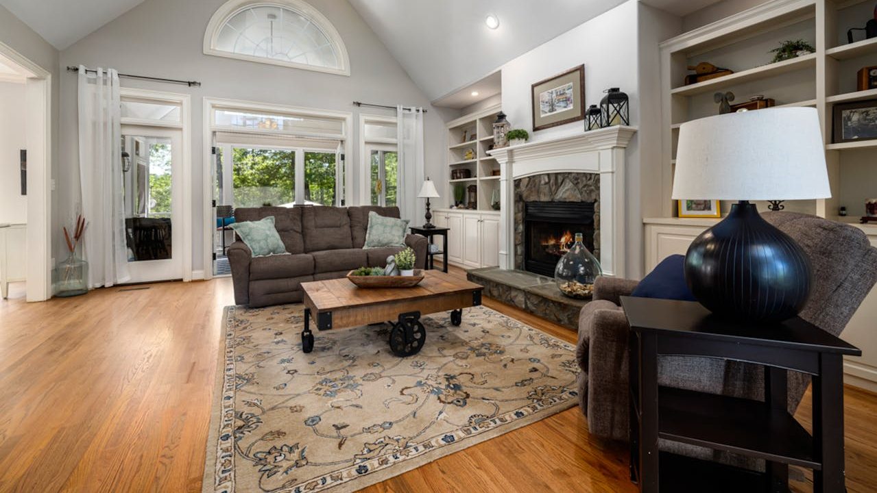 Cozy living room with high ceiling, fireplace, brown sofa, large windows, wooden floor, rug, and built-in shelves