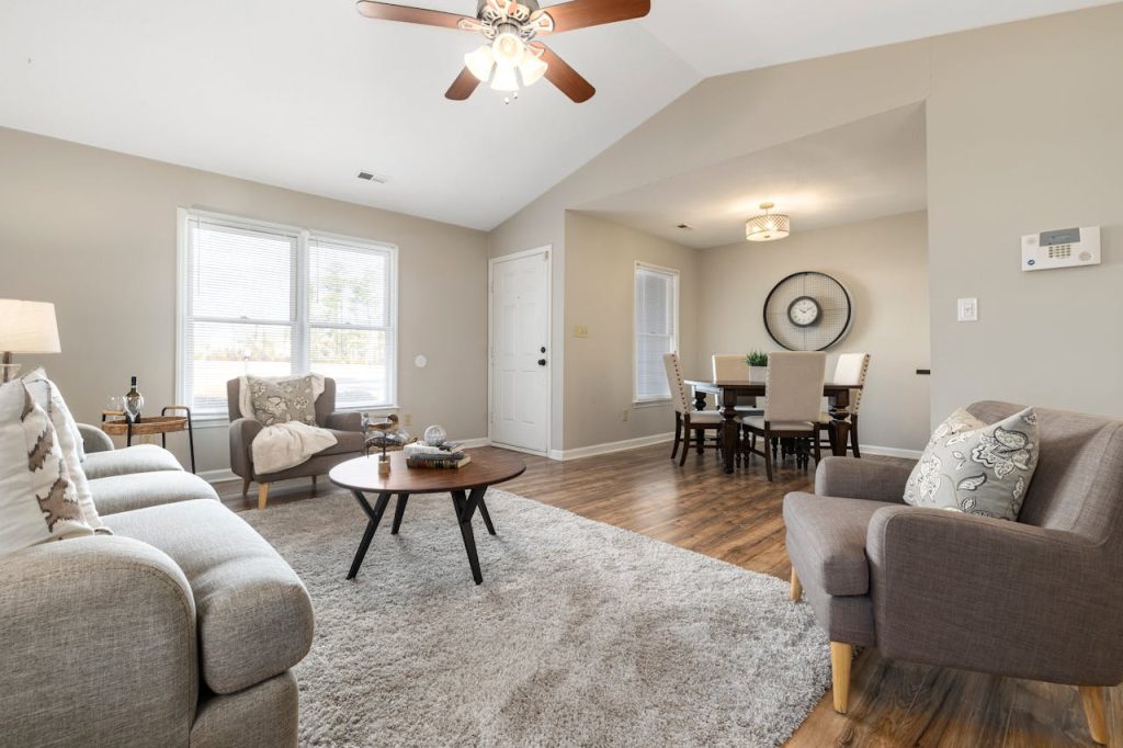 Cozy living and dining area with mid-century modern furniture, neutral tones, wood flooring, large windows, ceiling fan, and wall clock
