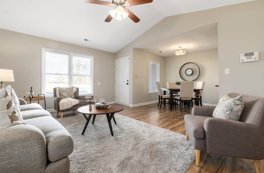 Cozy living and dining area with mid-century modern furniture, neutral tones, wood flooring, large windows, ceiling fan, and wall clock