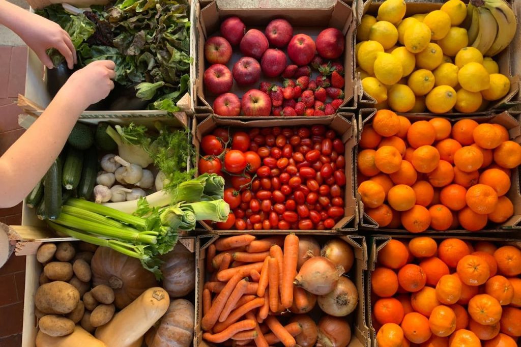 Cardboard boxes filled with fresh produce including vegetables, fruits, and leafy greens, with hands selecting items from left side