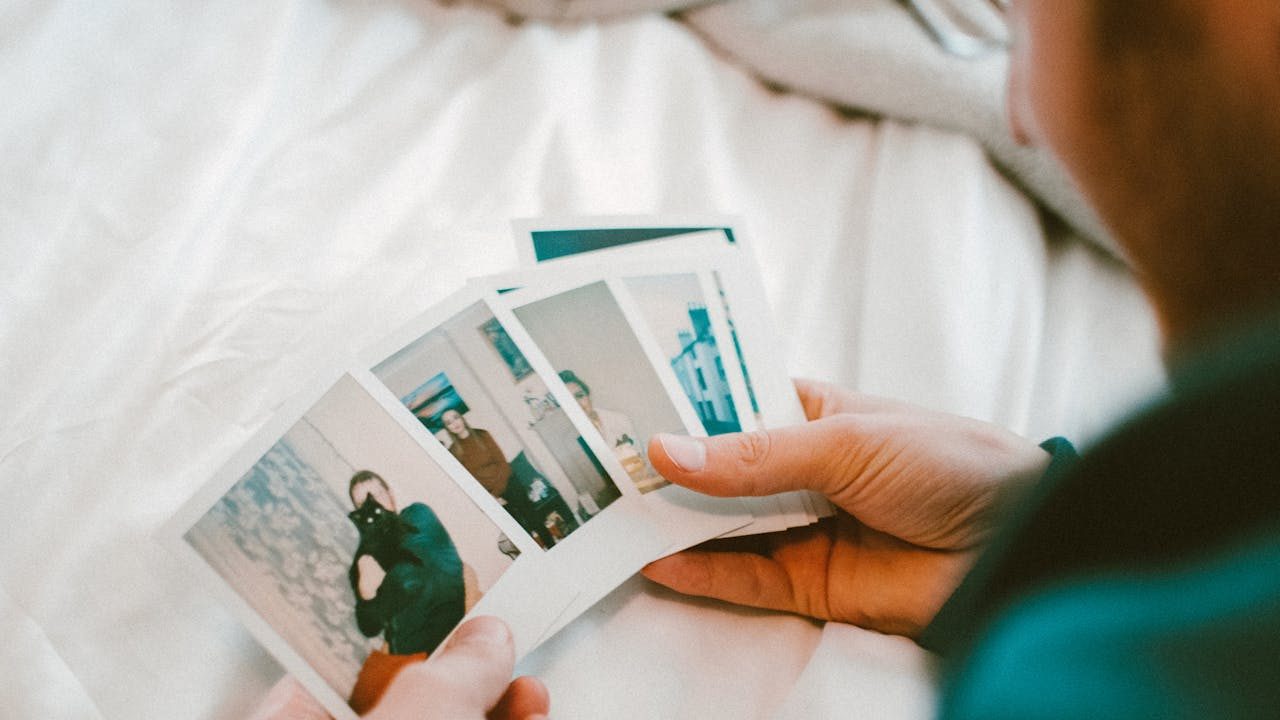 Person holding several instant photographs while sitting on a white bed, casually flipping through captured memories with both hands
