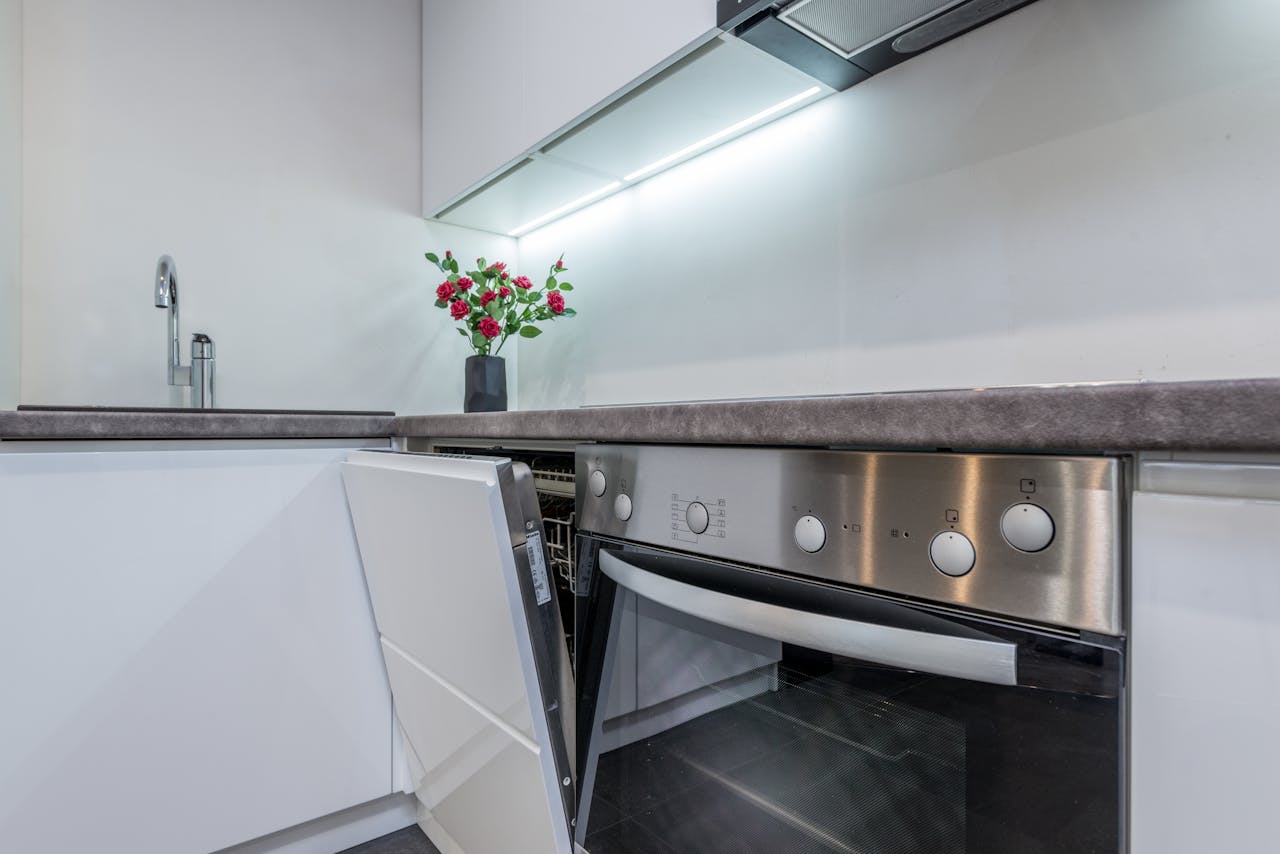 Modern kitchen with a sleek grey countertop, built-in stainless steel oven with the door open, integrated dishwasher slightly ajar, under-cabinet lighting, minimalist faucet