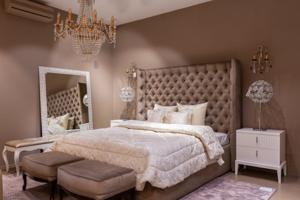 Luxurious bedroom with tufted headboard, chandelier, elegant bedding, mirrored decor, bedside tables, cushioned benches, and warm ambient lighting