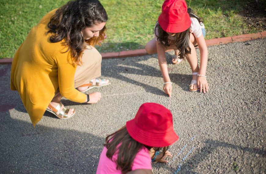 Three girls drawing on pavement with chalk in a park; girls wear red hats, it's sunny outside