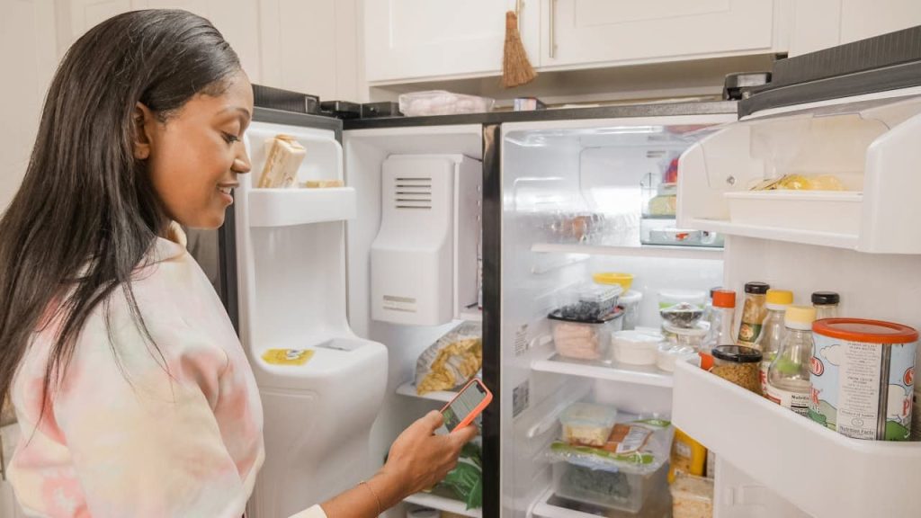 Woman with long dark hair looking at open refrigerator while checking her phone, wearing light-colored top in kitchen