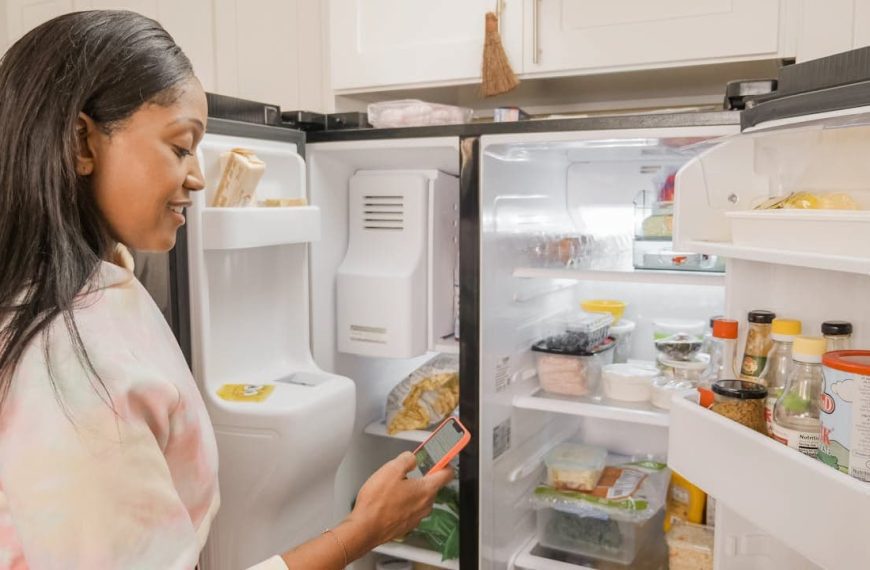 Woman with long dark hair looking at open refrigerator while checking her phone, wearing light-colored top in kitchen