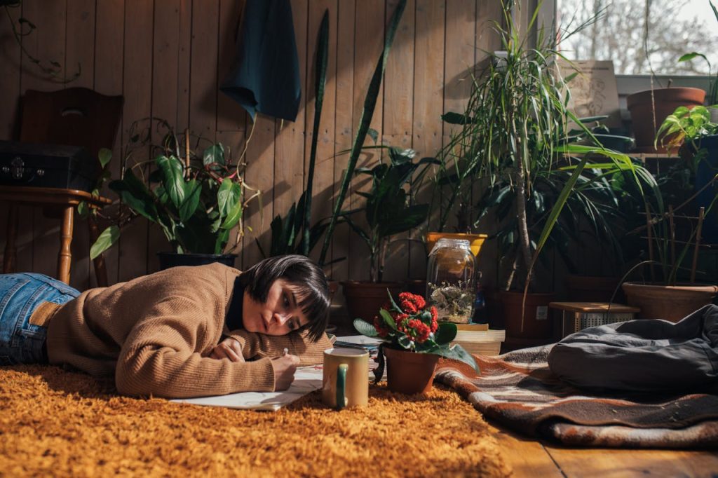 Person lying on patterned rug reading book, surrounded by houseplants and natural lighting from window
