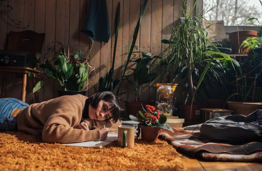 Person lying on patterned rug reading book, surrounded by houseplants and natural lighting from window