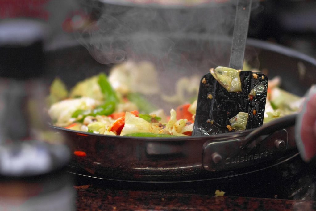 A steaming stir-fry of colorful vegetables cooks in a Cuisinart pan, stirred with a black spatula