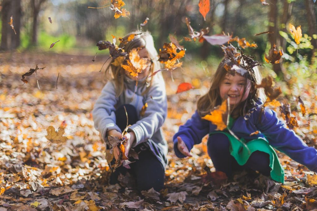 Two young children playing in autumn leaves, throwing colorful foliage in the air while crouching in a forest
