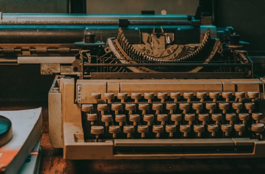Vintage mechanical typewriter with beige keys on wooden desk, displaying its internal mechanisms and roller, with books nearby