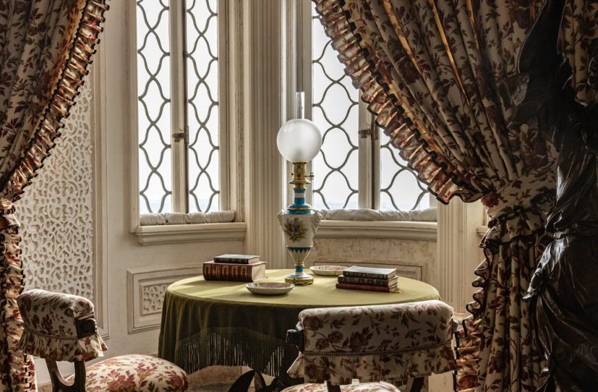 Vintage sitting area with floral chairs, round table, books, oil lamp, and ornate curtains near decorative windows