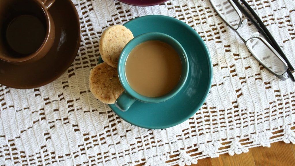 teal coffee cup filled with coffee on matching saucer, two cookies on the side, placed on a white crochet tablecloth