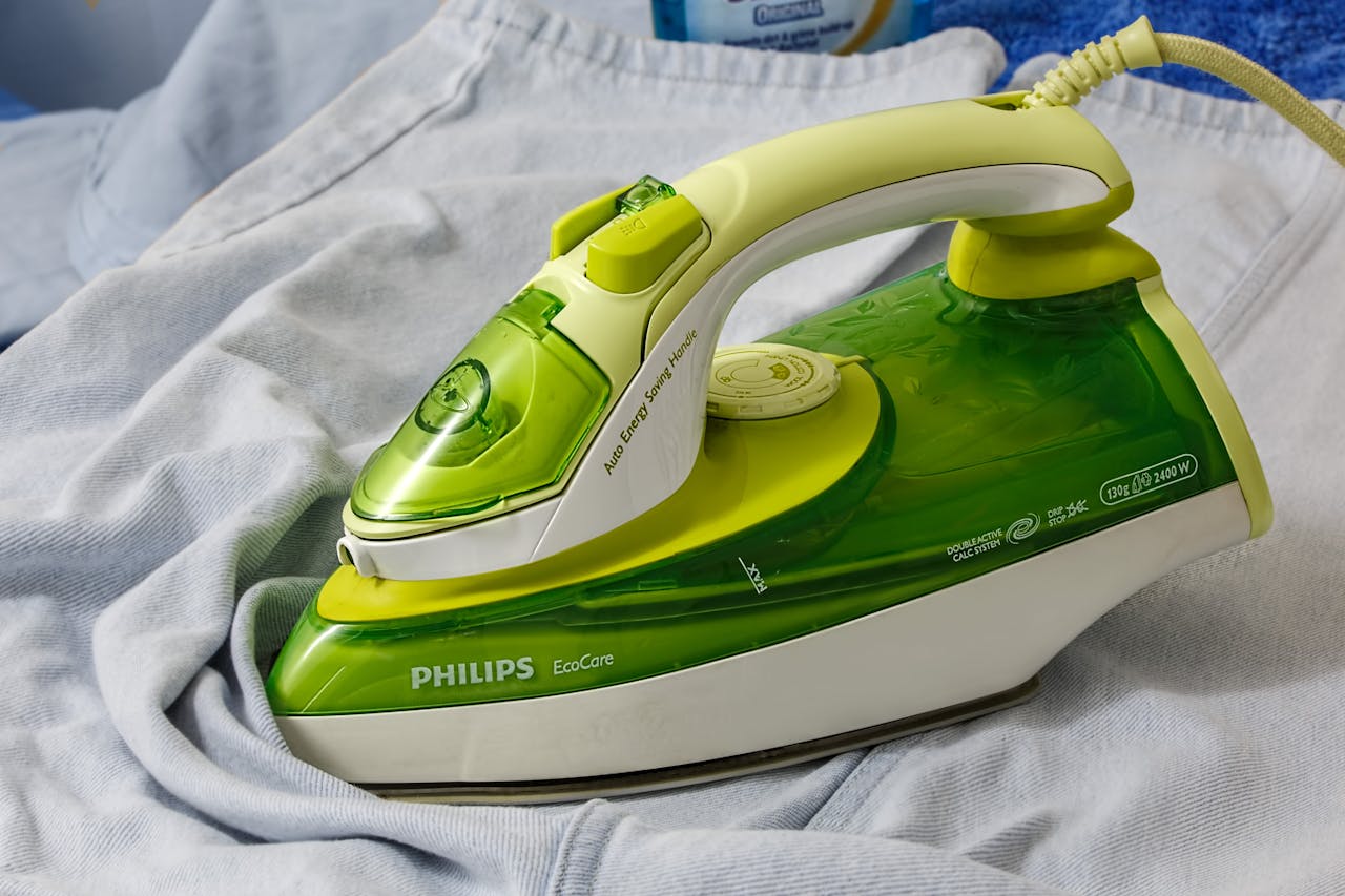 Close-up of a green and white Philips EcoCare steam iron placed on a wrinkled light blue shirt, featuring a translucent water tank, temperature dial, and energy-saving handle