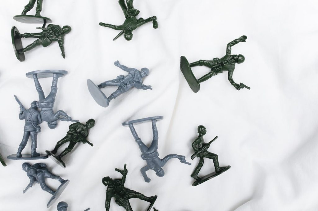 Plastic toy soldiers in green and gray colors scattered on white fabric, displaying various combat poses with rifles
