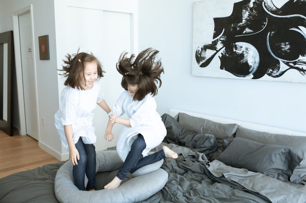 Two young girls in white tops and jeans joyfully jump on a messy bed in a modern, art-decorated bedroom