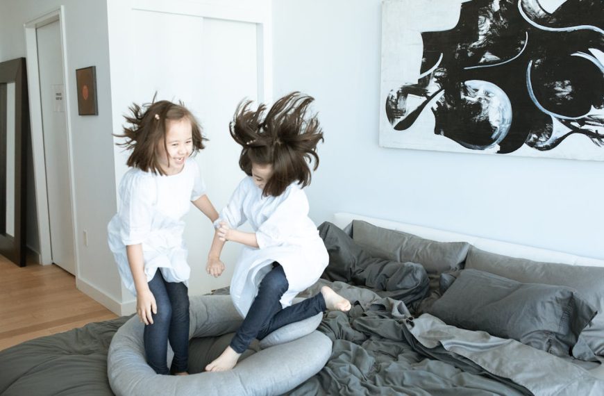 Two young girls in white tops and jeans joyfully jump on a messy bed in a modern, art-decorated bedroom