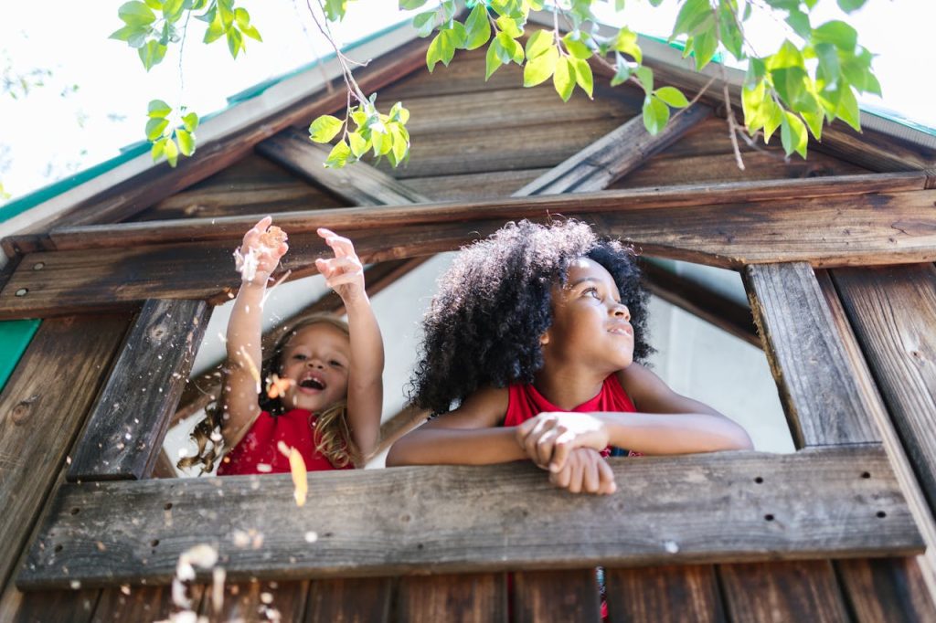 Two children play in a wooden treehouse, one leaning out the window, the other joyfully tossing leaves into the air