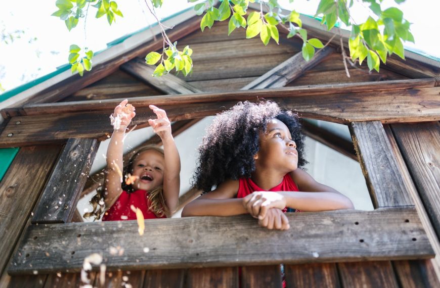 Two children play in a wooden treehouse, one leaning out the window, the other joyfully tossing leaves into the air
