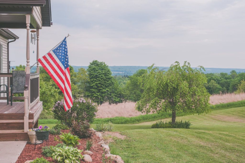 American flag displayed on rural house porch overlooking green landscape with trees, garden bed, and distant rolling hills