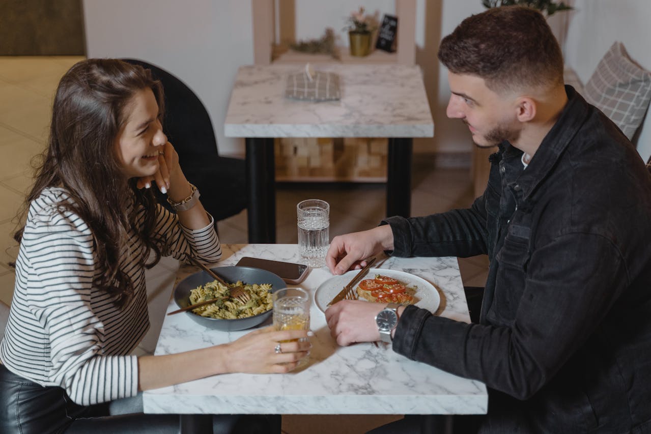 Young woman and man sitting at a marble table in a cozy restaurant, smiling and talking while eating pasta and toast, glasses of water and juice on the table, smartphone placed beside the plates