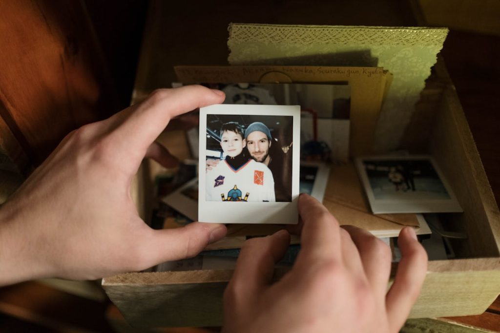 Hands holding a Polaroid photo of a man and child in hockey gear, surrounded by vintage photos and letters in a wooden box