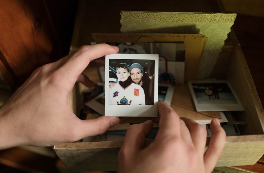 Hands holding a Polaroid photo of a man and child in hockey gear, surrounded by vintage photos and letters in a wooden box