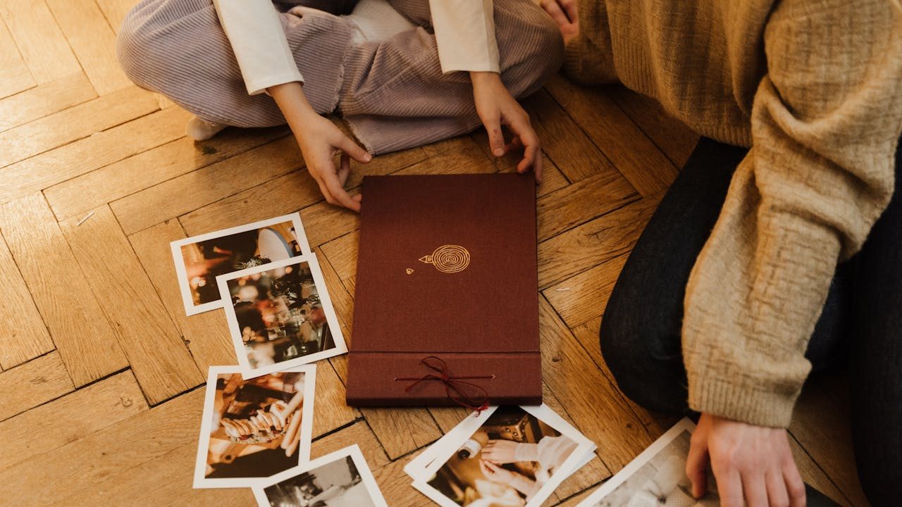 Two people sitting on a wooden floor, flipping through printed photographs, maroon photo album and loose pictures scattered