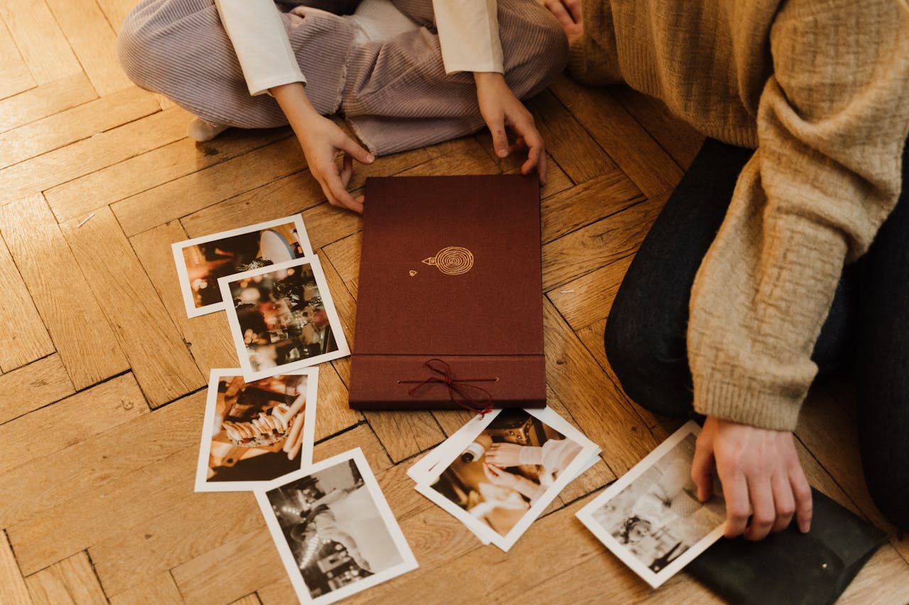 two people sitting on a wooden floor, flipping through printed photographs, maroon photo album and loose pictures scattered