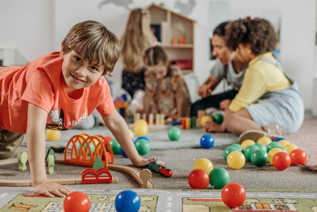 Smiling boy in red shirt playing on floor with toy train, colorful balls and wooden tracks around, children and adult in background