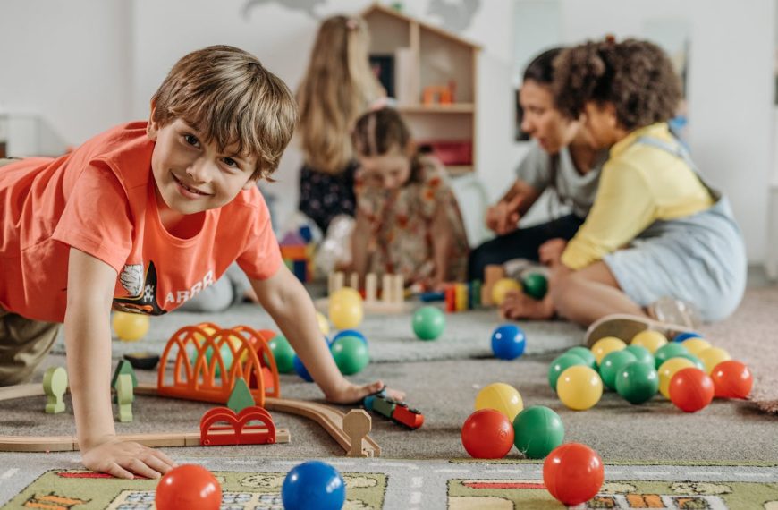 Smiling boy in red shirt playing on floor with toy train, colorful balls and wooden tracks around, children and adult in background